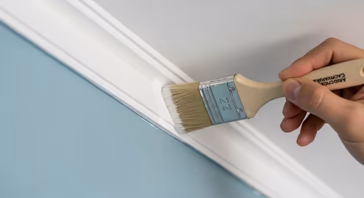 A steady hand using an angled brush to paint a clean, straight line where the white ceiling meets a colored wall.