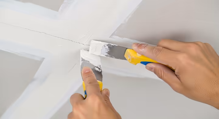 A close-up of a hand using a putty knife to apply spackling compound to a small crack in a drywall ceiling.
